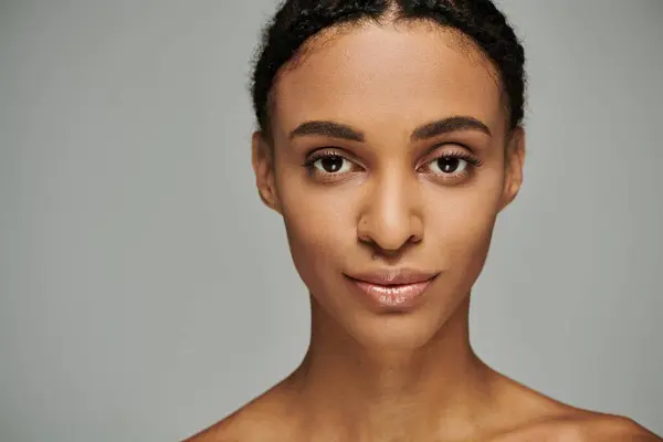A beautiful, young African American woman in a strapless top meticulously tending to her skin on a grey background. — Stock Photo