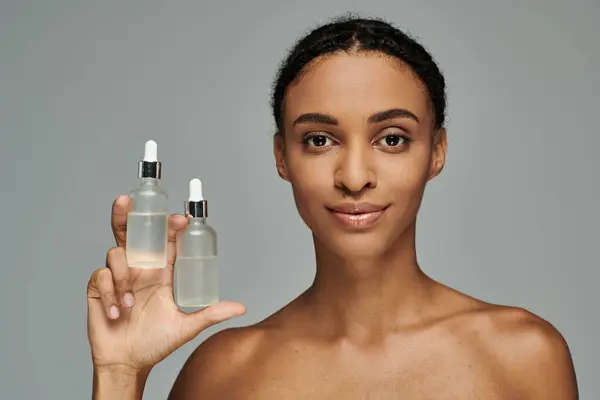 African American woman in strapless top holding up bottle of skin care product on grey background. — Stockfoto