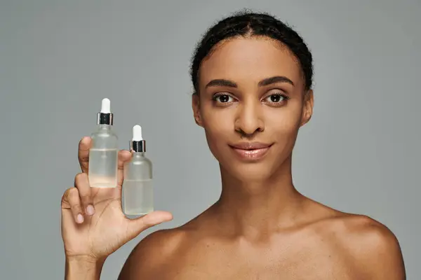 African American woman in strapless top holding up bottle of skin care product on grey background. — Stock Photo