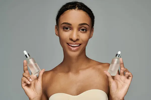 Young African American woman in strapless top taking care of skin, holding two serum bottles on grey background. — Stockfoto