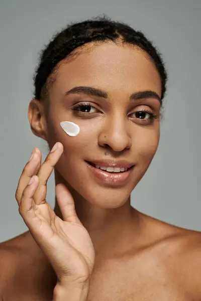 Junge Afroamerikanerin in schulterfreiem Top cremt ihr Gesicht vor grauem Hintergrund ein. — Stockfoto