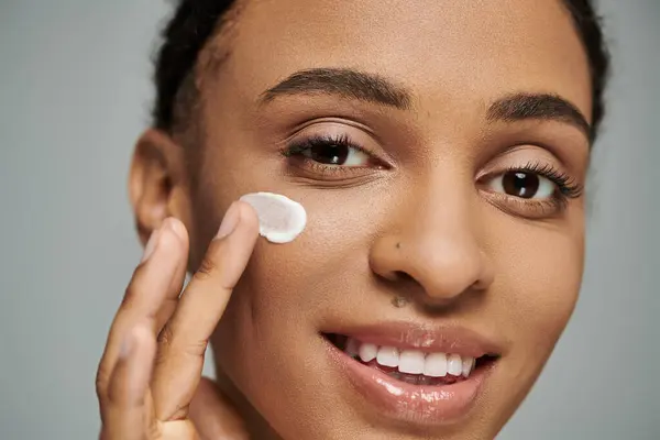 Jeune femme afro-américaine en haut sans bretelles, appliquant délicatement de la crème sur son visage sur un fond gris. — Photo de stock