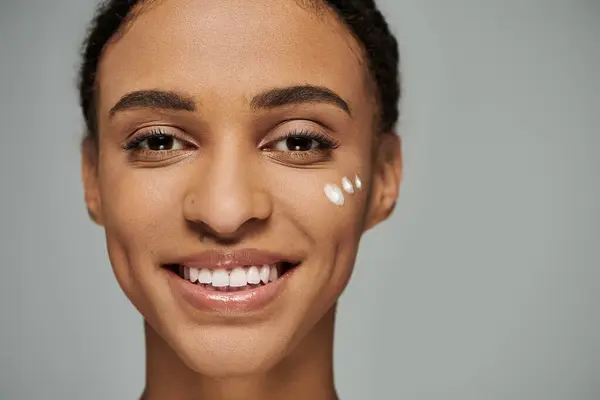 Young African American woman in strapless top, glowing with makeup, smiling on grey background. — Stock Photo