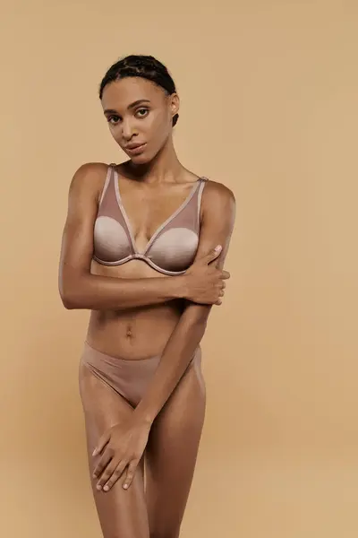 A pretty and slim African American woman in nude bra and panties, gracefully tending her body on a beige background. — Stock Photo