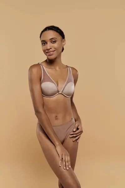 A slim African American woman gracefully poses in bra and panties, exuding confidence and self-care on a beige background. — Stock Photo
