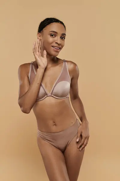Slim African American woman, donning a lingerie set, strikes a confident pose against a beige backdrop. — Stock Photo