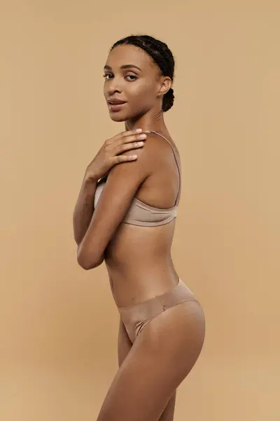 A pretty and slim African American woman in a tan bikini poses gracefully for the camera on a soft beige background. — Stock Photo
