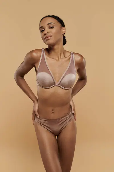 Elegante donna afroamericana in lingerie abbronzante, trasuda fiducia e si prende cura del suo corpo su uno sfondo beige. — Foto stock