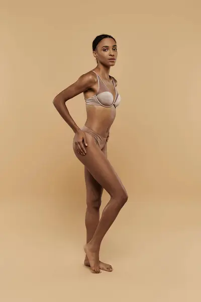 African American woman showcases her beauty in a bikini on a beige background, radiating confidence and grace. — Stock Photo