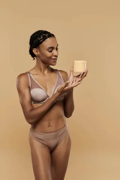 A slim African American woman in a bikini holding cream on a beige background. — Stock Photo
