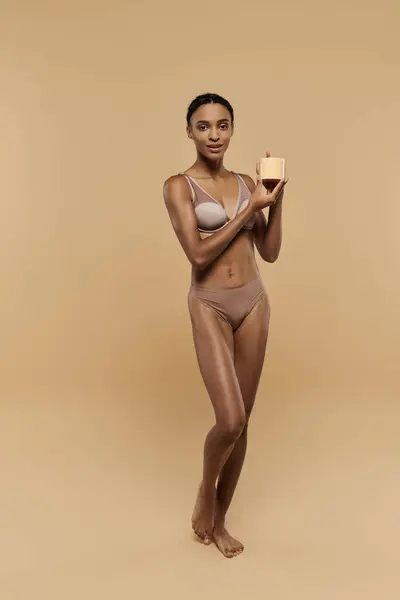 A stunning African American woman in a bikini gracefully holding cream a beige background. — Stock Photo