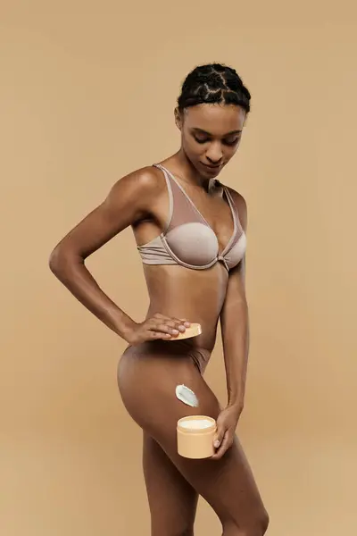 A stunning African American woman in a bikini delicately holds a jar of cream against a beige background. — Stock Photo