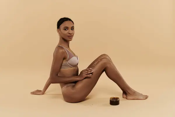 A pretty, slim African American woman in a bikini is sitting on the ground, taking care of her body in a serene setting. — Stock Photo
