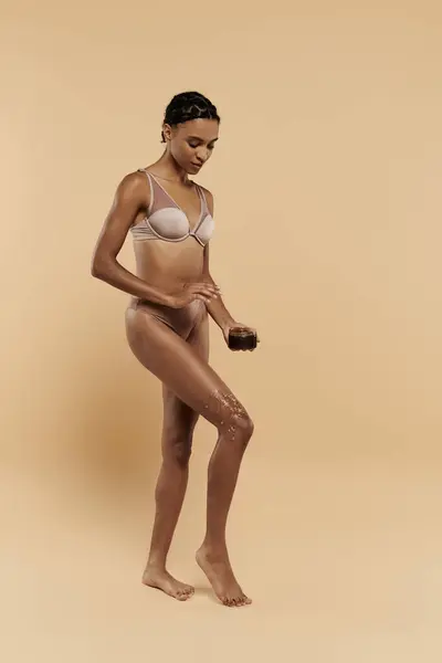 Una donna afroamericana magra e carina è in reggiseno bianco e mutandine abbronzanti, prendendosi cura del suo corpo su uno sfondo beige. — Foto stock
