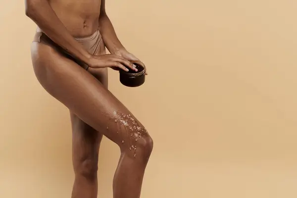 Una donna afroamericana elegante con una figura sottile posa con scrub caffè su gambe su uno sfondo beige. — Foto stock
