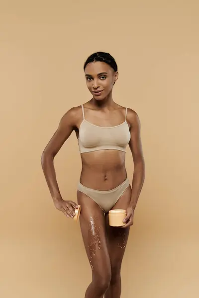 Una donna afroamericana magra in bikini in posa con scrub di zucchero sulle gambe su uno sfondo beige. — Foto stock