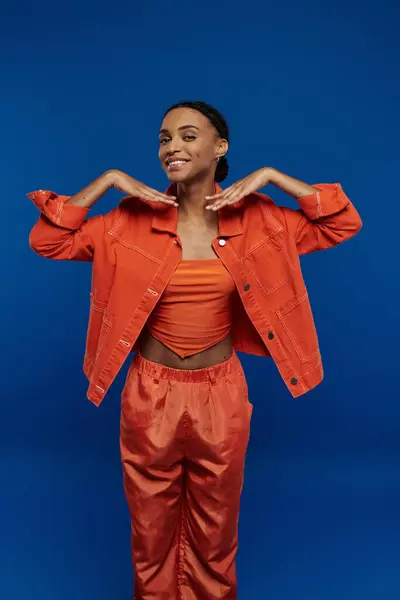 A pretty young African American woman stylishly dressed in an orange top and pants against a vibrant blue backdrop. — Stock Photo