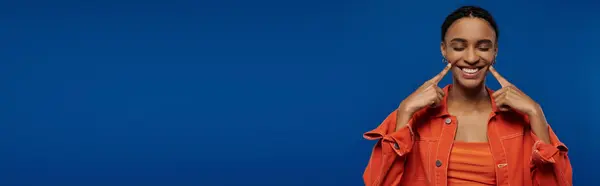 An African American man in an orange shirt makes a silly face against a blue backdrop. — Stockfoto