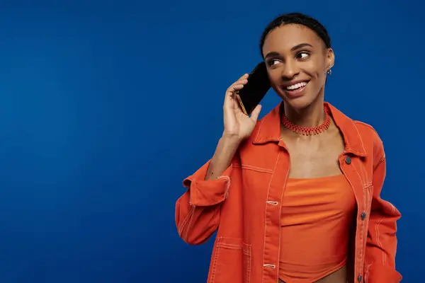 Une jeune femme afro-américaine élégante dans une chemise orange engagée dans un appel téléphonique sur un fond bleu vibrant. — Photo de stock