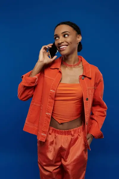 A young African American woman in a vivid orange outfit talks on a cell phone against a bold blue backdrop. — Stock Photo