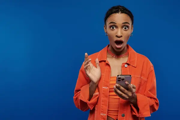 Hübsche junge Afroamerikanerin in orangefarbenem Hemd hält freudig ein Handy vor hellblauem Hintergrund. — Stockfoto