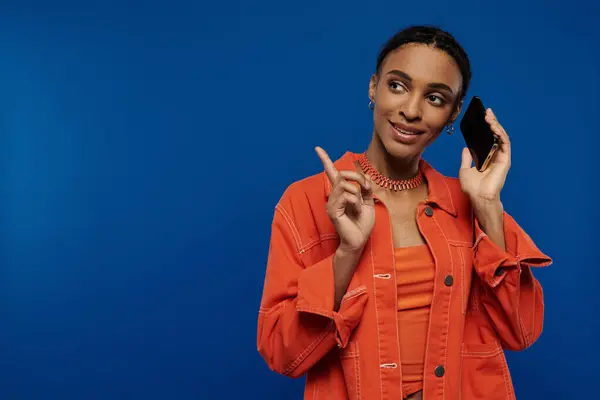 Junge Afroamerikanerin in leuchtend orangefarbenem Outfit mit einem Handy am Ohr vor blauem Hintergrund. — Stockfoto