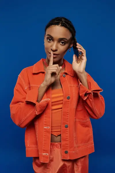 A pretty young African American woman standing confidently in a vibrant orange jacket and pants against a blue background. — Stock Photo