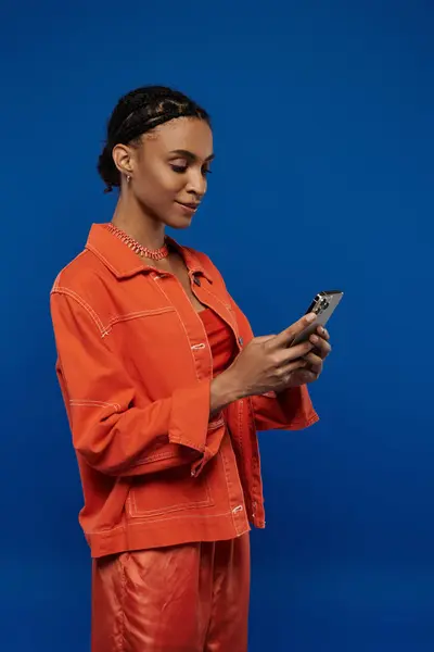 Junge Afroamerikanerin in leuchtend orangefarbenem Outfit mit Handy auf blauem Hintergrund. — Stockfoto