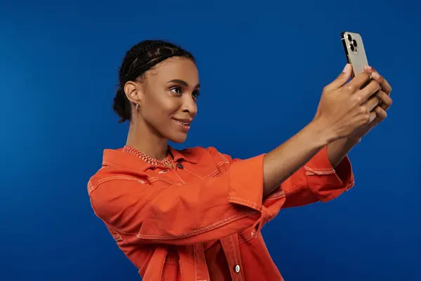 A young African American woman in vibrant orange outfit taking a picture with her cell phone against a blue background. — Stock Photo