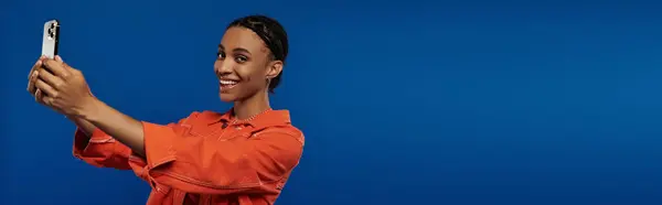 Vibrant young African American woman in orange outfit taking a picture with her cell phone against a bold blue backdrop. — Stock Photo