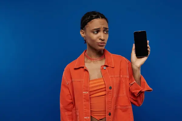 Una giovane donna afroamericana vestita di arancione regge un cellulare su uno sfondo blu. — Foto stock