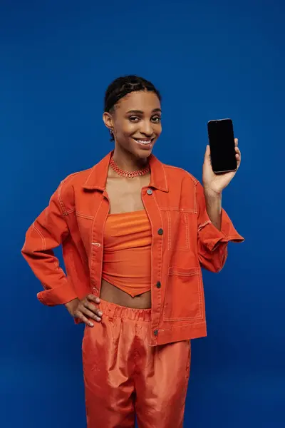 A stylish young African American woman in a bright orange outfit, holding a cell phone against a blue background. — Stock Photo