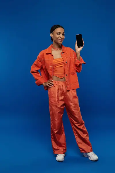 Eine junge Afroamerikanerin in leuchtend orangefarbenem Outfit hält ein Handy vor einem leuchtend blauen Hintergrund. — Stockfoto