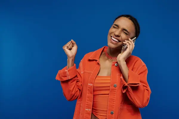 Uma jovem mulher afro-americana bonita com uma camisa laranja falando em um telefone celular contra um fundo azul. — Fotografia de Stock