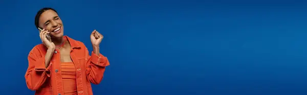 A pretty young African American woman in a vibrant orange shirt talking on a cell phone against a bright blue background. — Stock Photo