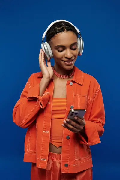 Joven mujer afroamericana en traje naranja, con auriculares, absorto en la pantalla del teléfono celular sobre fondo azul. - foto de stock