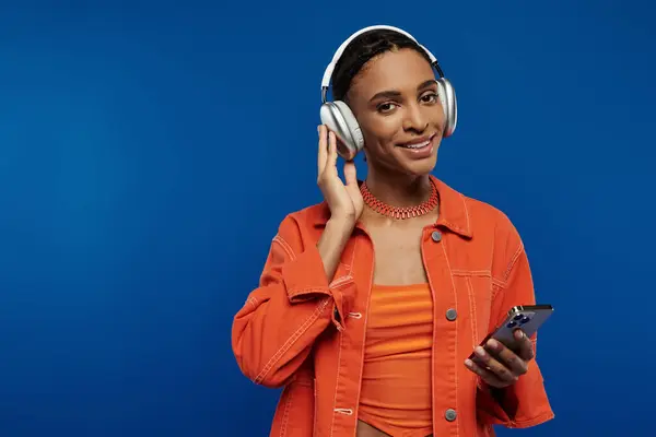 Young African American woman in vibrant orange outfit listening to music on headphones while holding a cell phone. — Stock Photo