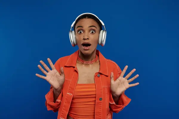 A stylish African American woman in an orange top listening to music on headphones against a blue background. — Stock Photo