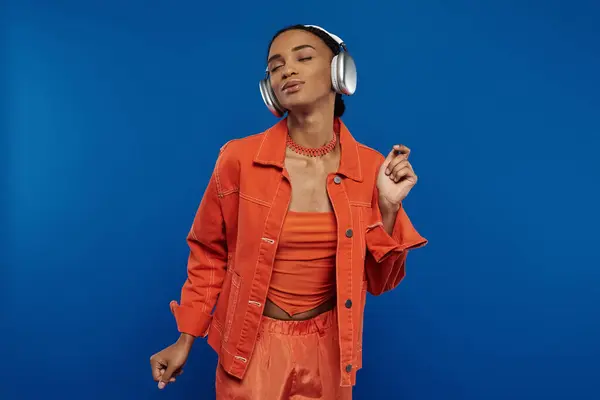 A young African American woman in a bright orange outfit listens to music with headphones against a blue background. — Stock Photo