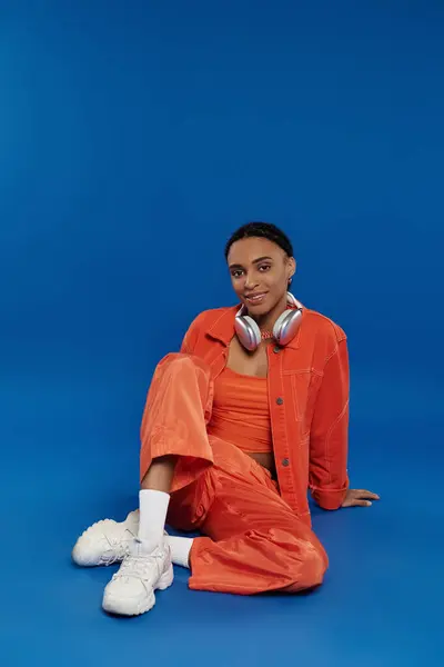 Eine junge Afroamerikanerin in leuchtend orangefarbenem Outfit sitzt auf dem Boden, durch Kopfhörer in Musik eingetaucht. — Stockfoto