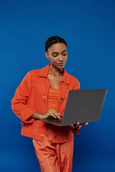 Une jeune Afro-Américaine en combinaison orange avec un ordinateur portable sur fond bleu. — Photo de stock