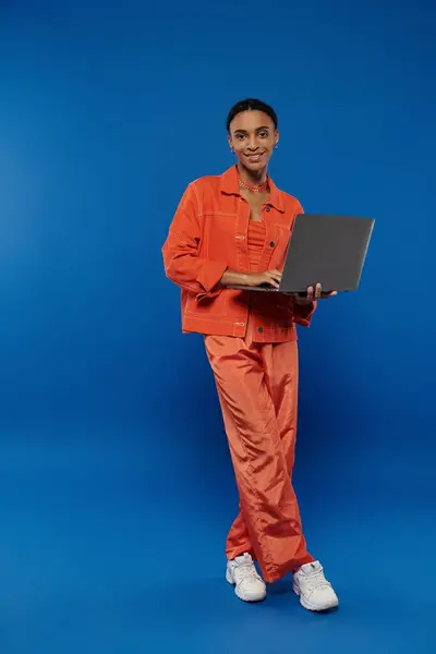 Giovane donna afroamericana in tuta arancione mette in mostra la creatività mentre tiene il computer portatile su uno sfondo blu vibrante. — Foto stock
