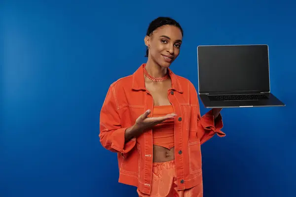 Una joven mujer afroamericana vibrante con una camisa naranja se centra en sostener una computadora portátil sobre un fondo azul. - foto de stock