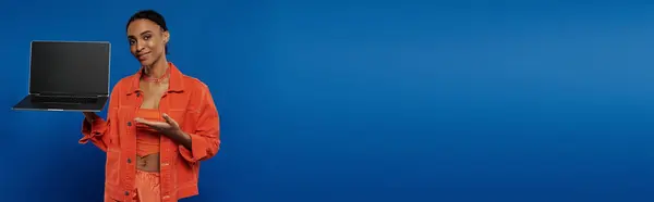 Young African American woman in vivid orange shirt actively using a laptop against a striking blue background. — Stock Photo