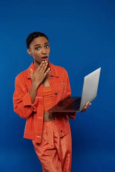 Young African American woman in orange outfit confidently holds a laptop against a vibrant blue backdrop. — Stock Photo