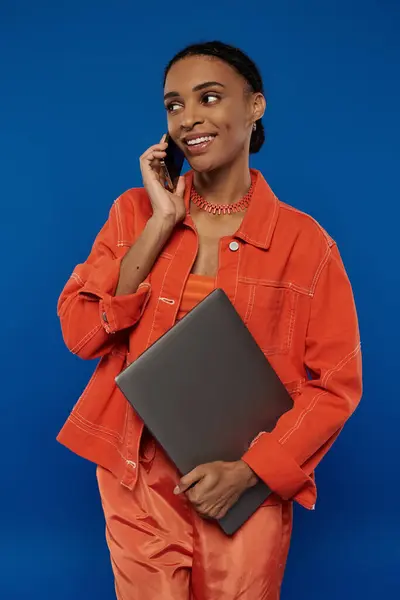 A young African American woman in a vibrant orange outfit talks on a cell phone while holding a laptop on a blue background. — Stock Photo