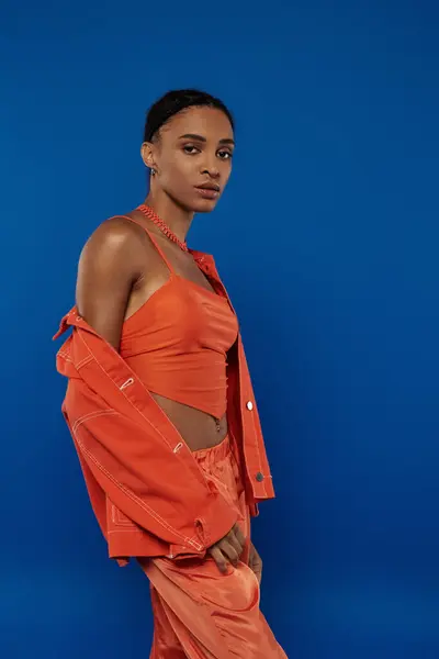 Pretty young African American woman in a vibrant orange outfit strikes a confident pose against a striking blue background. — Stock Photo