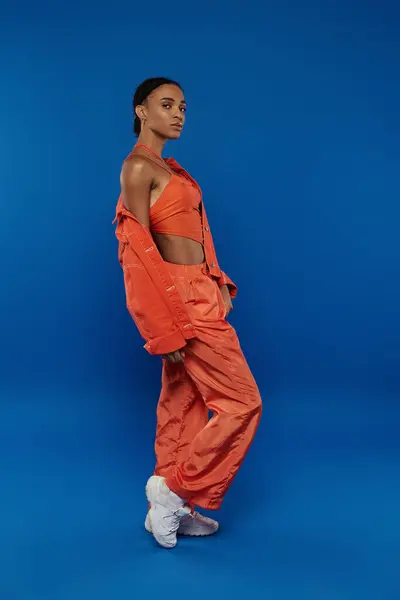 Une jolie jeune afro-américaine dans une tenue orange vibrante pose sur un fond bleu frappant. — Photo de stock