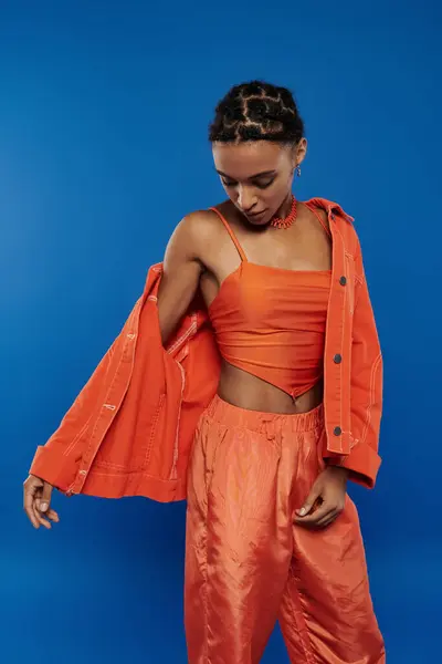 Eine hübsche junge Afroamerikanerin trägt selbstbewusst ein orangefarbenes Oberteil und eine Hose vor blauem Hintergrund. — Stockfoto
