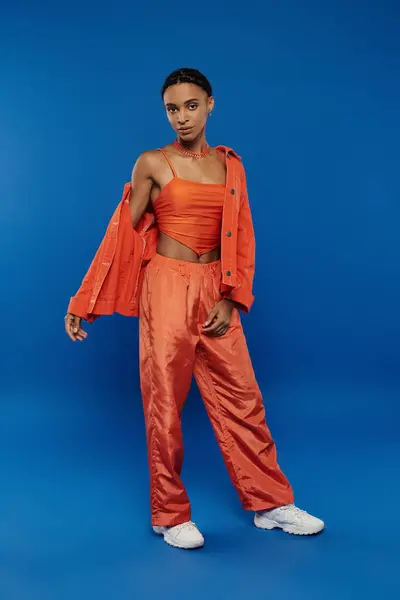 Eine hübsche junge Afroamerikanerin trägt ein orangefarbenes Oberteil und eine Hose auf blauem Hintergrund. — Stockfoto